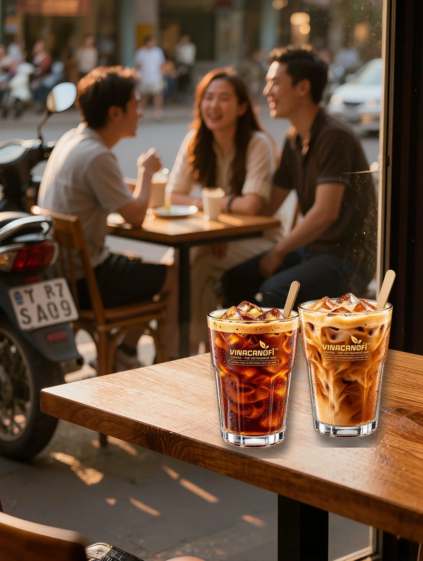 Hai ly cà phê đá trên bàn gỗ quán vỉa hè, phía sau là nhóm bạn trò chuyện và xe máy đỗ bên đường